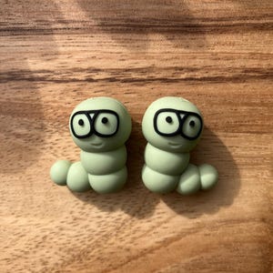Peut inclure: Deux jouets en silicone en forme de vers de couleur verte avec des yeux noirs et des lunettes. Les jouets sont sur une surface en bois.