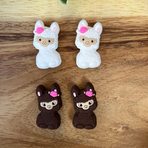 Peut inclure: Quatre petites figurines de lama, deux blanches et deux brunes. Chaque lama a une fleur rose sur la tête. Les lamas sont assis sur une surface en bois.