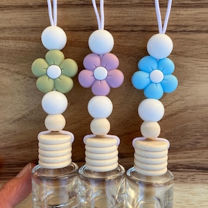 May include: Three car air fresheners with wooden beads and flower charms. The charms are green, purple, and blue. The air fresheners are hanging from white strings.