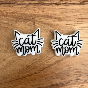 Peut inclure: Deux breloques blanches "maman chat" avec des contours noirs. Les breloques représentent un visage de chat avec le texte "maman chat" écrit à l'intérieur.