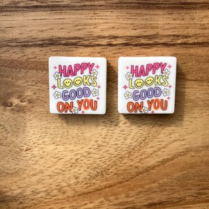 Peut inclure: Deux autocollants carrés blancs avec le texte "Happy looks good on you" en lettres roses, orange et jaunes avec de petits motifs de fleurs et de smileys.
