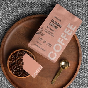 Puede incluir: Una bandeja de madera marrón con un tazón de granos de café, una tarjeta rosa con la palabra "COFFEE" impresa, una cuchara de metal dorada y una bolsa rosa de granos de café colombiano Supremo. La bolsa tiene el texto "COFFEE ROASTERS COLOMBIAN SUPREMO BOLD, RICH, NUTTY 100% ARABICA BEANS SOURCE: HUILA, COLOMBIA" impreso.