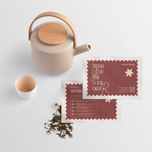 Könnte beinhalten: Eine beige Keramik-Teekanne mit Holzgriff und Deckel, eine weiße Teetasse und zwei weinrote Karten mit dem Text "Thank You For Your Order". Lose Teeblätter sind in der Nähe verstreut. Die Karten haben ein Blumendesign.