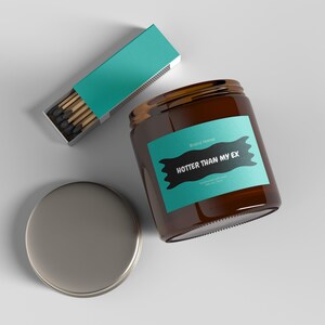 Op de afbeelding: Een bruine glazen potkaars met een turquoise label met de tekst "HOTTER THAN MY EX". Een zilveren deksel ligt naast de pot en een turquoise lucifersdoos is in de buurt geopend.
