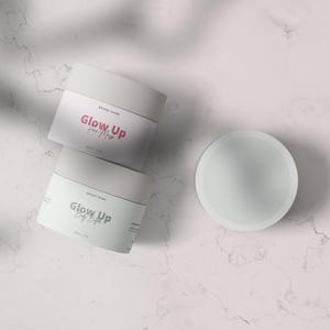 May include: Three white cosmetic jars with the text "Glow Up" in pink and green, and a separate lid. The jars are stacked on a marble surface, with a shadow cast across the scene. The product is labeled "Face Mask" and "Daily Brights".