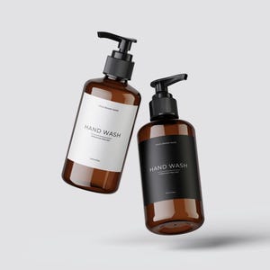 Puede incluir: Dos botellas de vidrio ámbar con tapas de bomba negras y etiquetas que dicen "HAND WASH" en texto blanco y negro. Las botellas flotan en el aire sobre un fondo blanco.