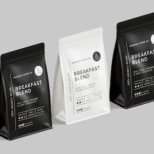 Op de afbeelding: Drie koffiezakken, twee zwarte en één witte, worden getoond. De zakken zijn voorzien van het opschrift "Breakfast Blend" met details over het smaakprofiel van de koffie. De zakken bevatten ook de tekst "100% Arabica Beans."