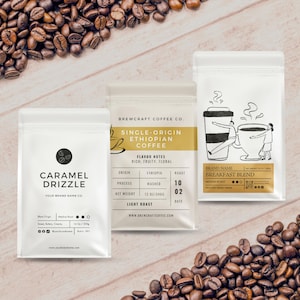 May include: Three white coffee bags with different labels and coffee beans scattered around. The bags are labeled "Caramel Drizzle", "Single-Origin Ethiopian Coffee", and "Breakfast Blend". The bags are filled with coffee.