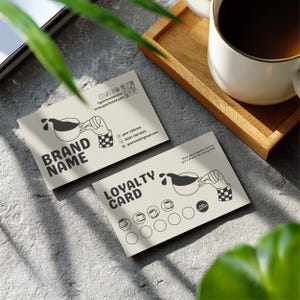 May include: Two beige coffee shop loyalty cards with a coffee cup graphic and the words "BRAND NAME" and "LOYALTY CARD". A white mug of coffee sits on a wooden tray. The cards have contact information and a QR code.