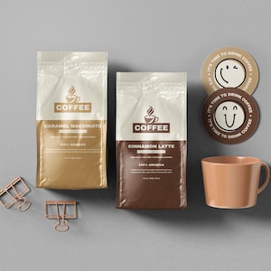 Op de afbeelding: Overzichtsfoto van koffieproducten. Twee verzegelde koffiezakken, één goudkleurig en één bruin, zijn gelabeld met "Caramel Macchiato" en "Cinnamon Latte". Twee ronde onderzetters met smiley-gezichten en de tekst "It's time to drink coffee" zijn aanwezig, samen met een perzikkleurige mok.
