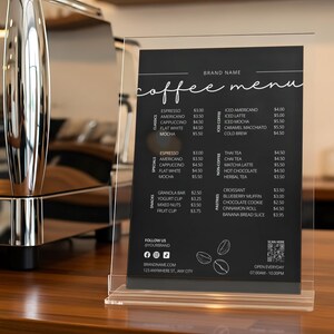 Può includere: Un menu di caffè nero esposto in un supporto in acrilico trasparente. Il menu elenca varie bevande al caffè, tè e pasticcini. Il menu include il nome del marchio e le informazioni di contatto.