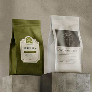 May include: Two tea packages on a gray surface. One is olive green with "Herbal Tea" on the label. The other is white with "Chamomile Tea" on the label. Both packages are standing upright.