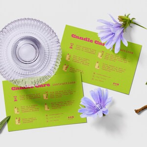 May include: A clear glass candle holder with a ridged design sits atop two green cards with candle care instructions. The cards are decorated with illustrations and text in pink and white. Purple flowers and greenery are scattered around the cards.