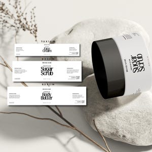 Peut inclure: Un pot blanc de gommage au sucre avec un couvercle noir, accompagné de trois étiquettes rectangulaires. Les étiquettes mesurent 19,05 x 3,81 cm pour la crème visage, 20,32 x 6,35 cm pour le gommage et 21,59 x 5,08 cm pour le beurre corporel.