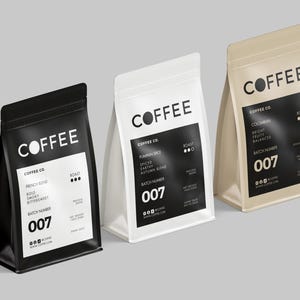 May include: Three coffee bags in black, white, and beige. Each bag features the word "COFFEE" in white, with a black label detailing the coffee blend, roast, and batch number. The bags are standing on a grey surface.