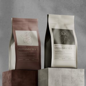 Könnte beinhalten: Zwei Teepackungen, eine braune und eine cremefarbene, werden vor einem grauen Hintergrund gezeigt. Die braune Packung trägt die Aufschrift "Peppermint Tea" mit einer botanischen Illustration. Die cremefarbene Packung trägt die Aufschrift "Chamomile Tea" mit einer floralen Illustration.