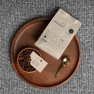 Op de afbeelding: Een bruine papieren zak met de tekst "100% ARABICA ETHIOPIAN COFFEE CO" erop gedrukt. De zak bevat koffiebonen en informatie over de herkomst, de branding en de branddatum. De zak ligt op een houten dienblad met een kom vol koffiebonen en een gouden lepel.