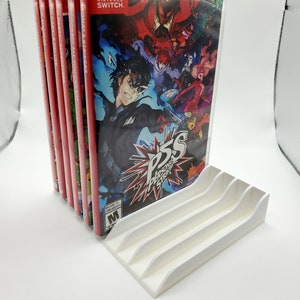 May include: A white plastic stand with four slots for storing Nintendo Switch game cartridges. The stand is holding a stack of six game cartridges, including "Persona 5 Strikers".