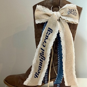 May include: Brown leather cowboy boot adorned with a beige bow and ribbons. The bow features the text "Luke Combs" and the ribbon says "Beautiful Crazy" in navy blue. The boot has a decorative design.