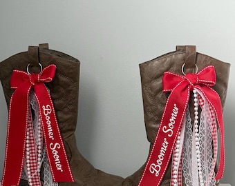 May include: A pair of brown cowboy boots with decorative red bows and ribbons. The ribbons feature the text "Boomer Sooner", along with white lace and checkered accents. The boots are displayed on a white surface.