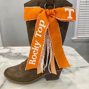 Op de afbeelding: Bruine leren cowboylaars met een oranje lint en de tekst "Rocky Top" in het wit. Het lint is geaccentueerd met wit kant en een ruitpatroon. De letter "T" is ook zichtbaar op het lint.