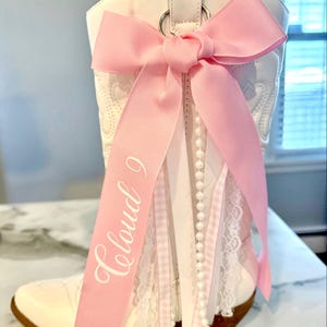 Peut inclure: Une botte de cowboy blanche ornée d'un grand nœud rose et de rubans décoratifs. La botte a une semelle marron et est ornée d'un ruban rose avec l'inscription "Cloud 9". De la dentelle blanche et un rang de perles complètent la décoration.