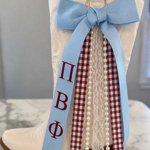 May include: A white western-style boot adorned with a light blue ribbon bow featuring the Greek letters Pi Beta Phi in dark red. The decoration includes two red and white gingham ribbons, a white lace trim, and two strands of white faux pearls.