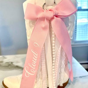 May include: A white western-style boot adorned with a large light pink ribbon bow attached to a silver ring. The ribbon features the text Cloud 9 in white script. Additional decorative elements include a string of white pearls and white lace trim.