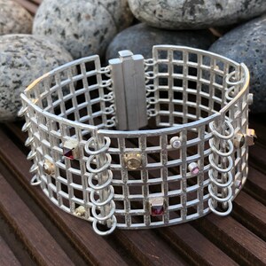 May include: A silver bracelet with a grid pattern and various colored gemstones. The bracelet has a hinged clasp and is made of sterling silver.