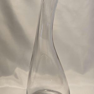 May include: A clear glass decanter featuring a wide, rounded base that tapers into a tall, slender, and slightly curved neck. The vessel has a smooth, transparent surface and a thick, solid glass bottom, showcasing a simple and functional design.