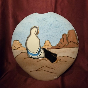 May include: A ceramic wall hanging with a Southwestern desert scene. The artwork depicts a figure in a white robe with a blue stripe, seated and facing away from the viewer. The background features mesas under a light blue sky and a tan desert floor.