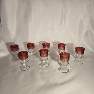 May include: A set of eight vintage wine glasses. Each glass features a clear, textured base and a ruby-red bowl. The glasses are arranged on a white surface, showcasing their elegant design and color contrast.