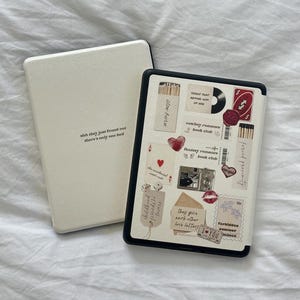 Puede incluir: Una funda blanca para lector de libros electrónicos con borde negro y tapa a juego. La tapa tiene un collage de pegatinas, incluyendo un disco, corazones y texto. El texto en la tapa dice "shh they just found out there's only one bed."