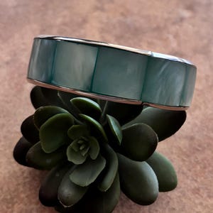 May include: A wide bangle bracelet featuring a series of rectangular, iridescent teal-colored shell inlays set within a polished silver-toned metal frame. The bracelet has a smooth, reflective surface with visible natural grain patterns in the shell.
