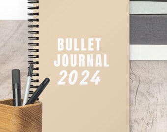 May include: A beige spiral bound journal with the text "BULLET JOURNAL 2024" printed in white on the front cover.