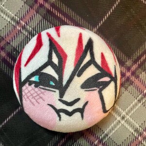 Puede incluir: Un bot&oacute;n redondo y suave de color blanco con un dise&ntilde;o de rostro estilizado. La cara presenta contornos negros, acentos rojos y mejillas rosadas. Los ojos son de color azul claro. El bot&oacute;n est&aacute; sobre un fondo a cuadros.