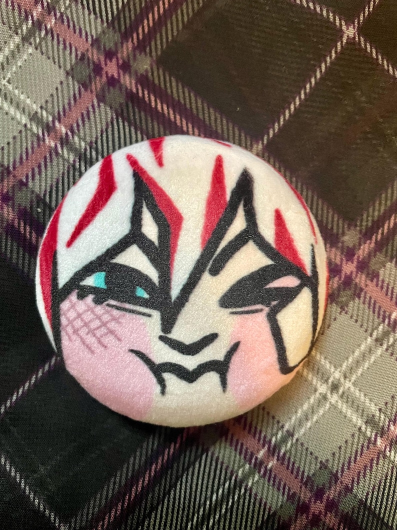 Puede incluir: Un bot&oacute;n redondo y suave de color blanco con un dise&ntilde;o de rostro estilizado. La cara presenta contornos negros, acentos rojos y mejillas rosadas. Los ojos son de color azul claro. El bot&oacute;n est&aacute; sobre un fondo a cuadros.