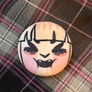 Puede incluir: Un bot&oacute;n redondo con un dise&ntilde;o de cara de dibujos animados. La cara tiene contornos negros, mejillas rosadas y una boca negra con colmillos. El fondo es un patr&oacute;n de cuadros en tonos grises, negros y morados.