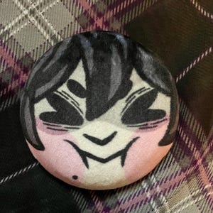 Puede incluir: Un peluche redondo con una cara de estilo anime. Tiene pelo, ojos y boca negros, y mejillas rosas. El peluche est&aacute; sobre un fondo a cuadros con tonos grises, blancos y morados.