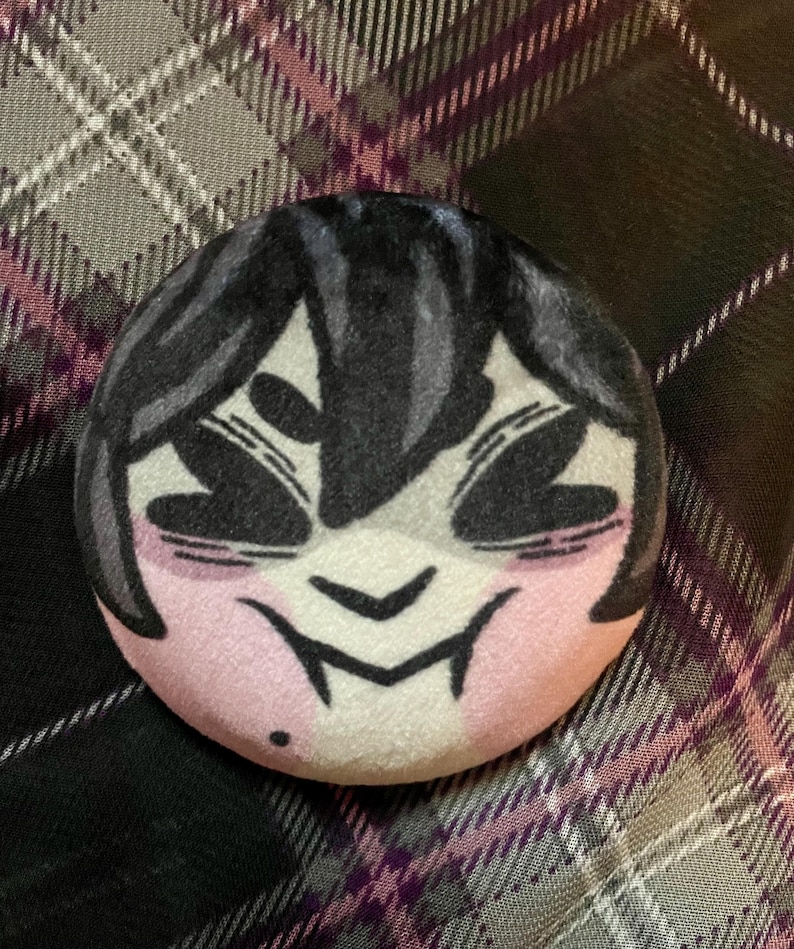 Puede incluir: Un peluche redondo con una cara de estilo anime. Tiene pelo, ojos y boca negros, y mejillas rosas. El peluche est&aacute; sobre un fondo a cuadros con tonos grises, blancos y morados.