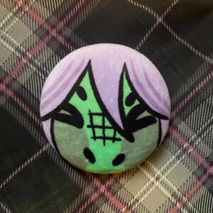 Puede incluir: Un bot&oacute;n redondo con un dise&ntilde;o de dibujos animados. Presenta una cara verde con detalles negros, cabello morado y un patr&oacute;n de cuadr&iacute;cula. El fondo es un patr&oacute;n de cuadros en tonos grises, rosas y negros.