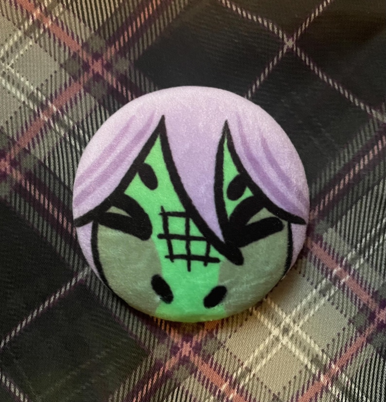 Puede incluir: Un bot&oacute;n redondo con un dise&ntilde;o de dibujos animados. Presenta una cara verde con detalles negros, cabello morado y un patr&oacute;n de cuadr&iacute;cula. El fondo es un patr&oacute;n de cuadros en tonos grises, rosas y negros.