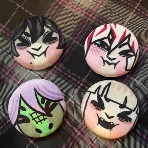 Puede incluir: Cuatro botones redondos con caras de dibujos animados. Cada bot&oacute;n presenta un personaje diferente con cabello y expresiones faciales &uacute;nicos. La paleta de colores incluye rosa, negro, blanco, verde y morado. Los botones est&aacute;n sobre un fondo a cuadros.