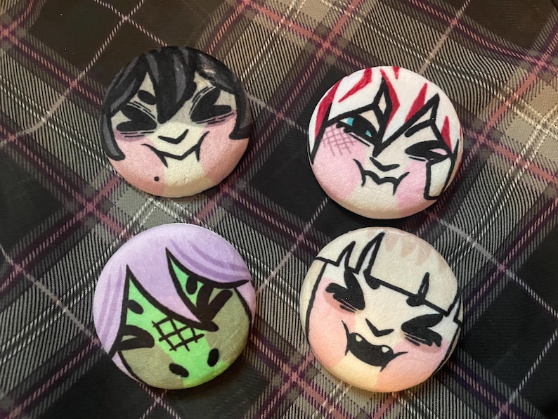 Puede incluir: Cuatro botones redondos con caras de dibujos animados. Cada bot&oacute;n presenta un personaje diferente con cabello y expresiones faciales &uacute;nicos. La paleta de colores incluye rosa, negro, blanco, verde y morado. Los botones est&aacute;n sobre un fondo a cuadros.