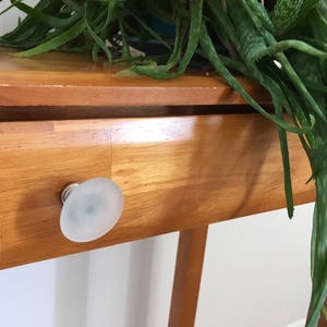May include: A wooden end table with a single drawer and a frosted glass knob. The table is light brown and has a natural wood finish. A green plant is hanging over the table.