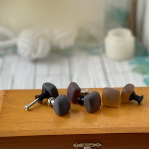 May include: Five square-shaped cabinet knobs with a frosted glass finish. The knobs are in shades of brown, gray, and clear. The knobs are arranged on a wooden surface.