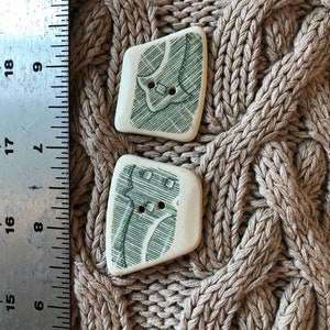 May include: Two ceramic buttons with a green and white design. The buttons are shaped like a rectangle with rounded corners. The design on the buttons is a stylized image of a building.