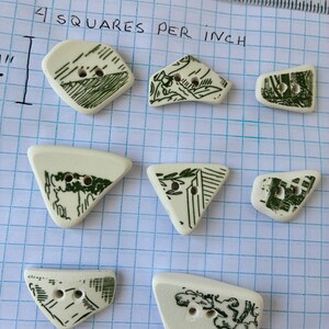 May include: A collection of eight irregularly shaped, off-white ceramic buttons with two holes each. Each button features a unique green illustration. The text "4 squares per inch" is handwritten at the top of the image.