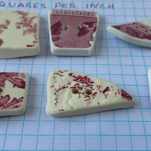 May include: A collection of ceramic buttons in various shapes, featuring a cream base with a burgundy floral pattern. The buttons have two holes for sewing and are arranged on a grid background.