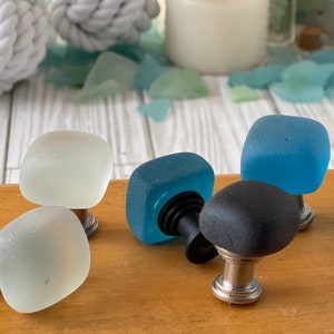 May include: Four square cabinet knobs with frosted glass tops in white, blue, and black. The knobs have a silver metal base.